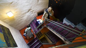 Une personne descend un escalier dont les marches sont violettes et les rampes en métal, dans une pièce dont le mur est blanc et présente une texture particulière, et qui est ornée d’une fresque colorée.