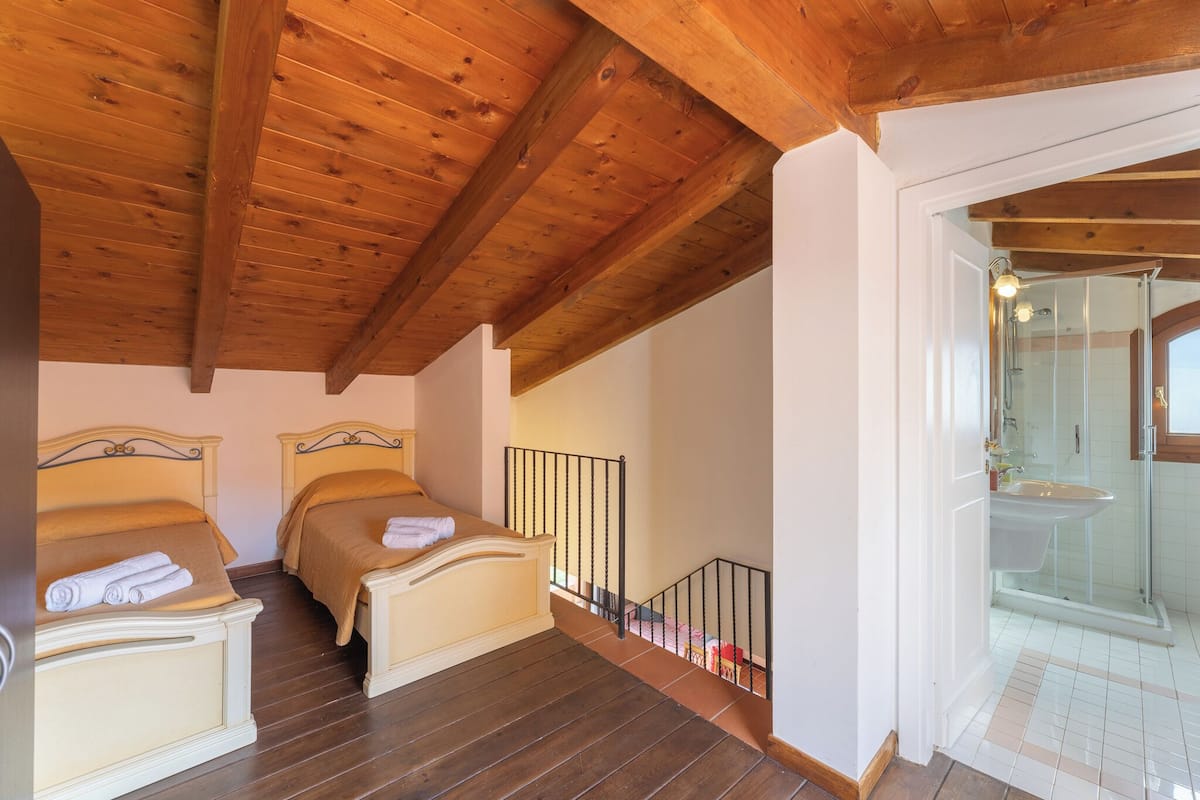 A bedroom with two beds, wooden ceiling beams, a small balcony, and a bathroom with a glass shower.