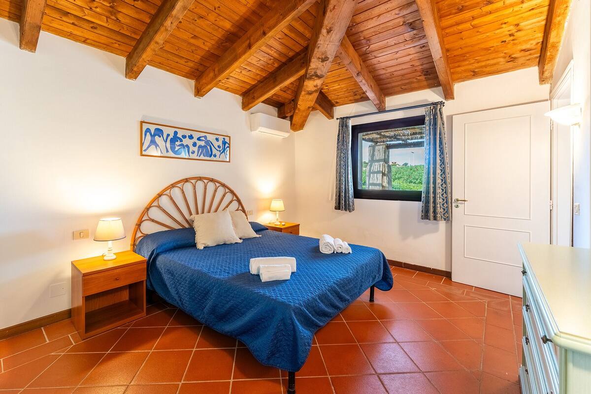 A bedroom with a wooden ceiling, a bed with blue bedding, two bedside lamps, a window with curtains, and a door.