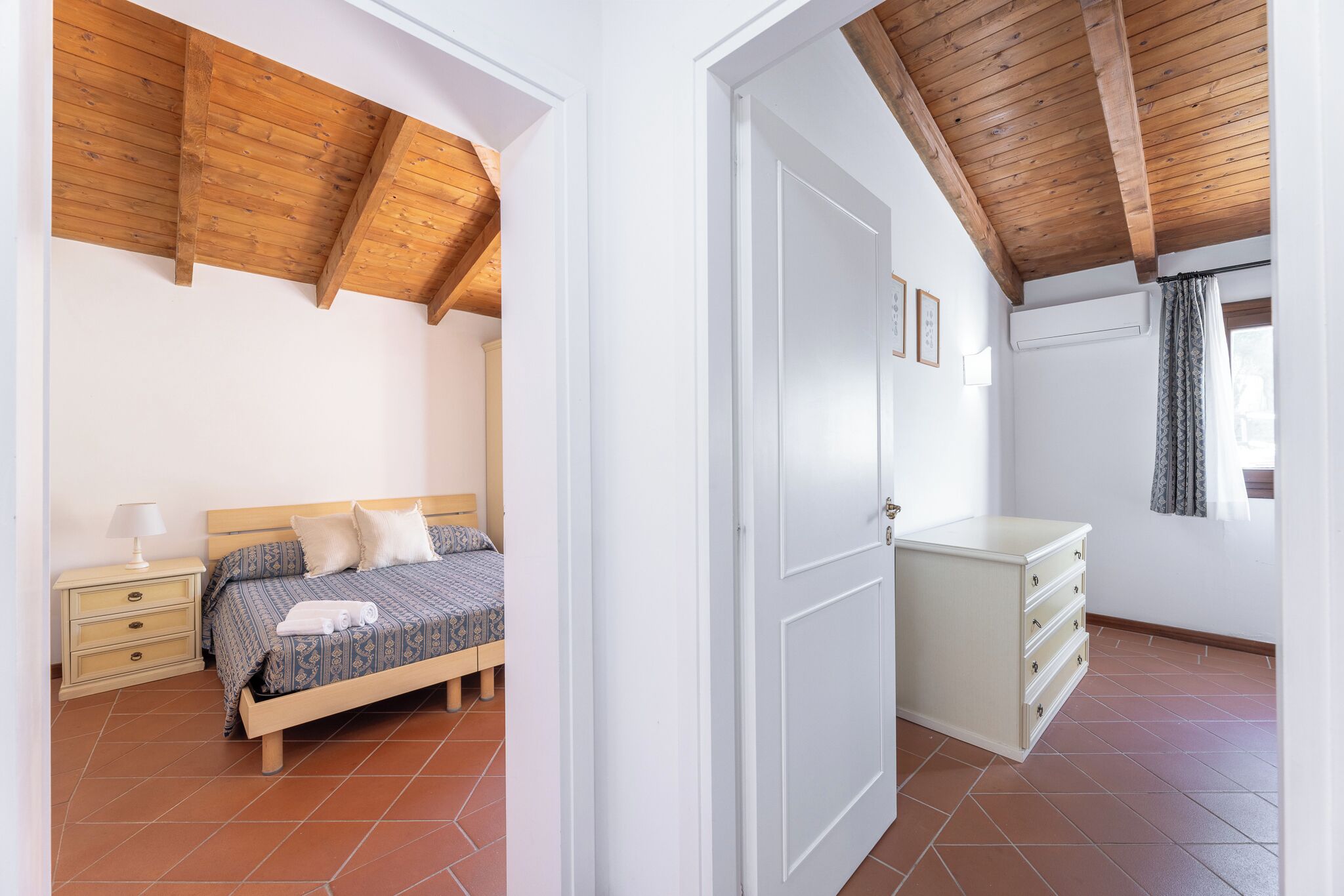 A bedroom with a wooden ceiling, a bed with a patterned bedspread, a bedside table with a lamp, and a dresser with drawers.