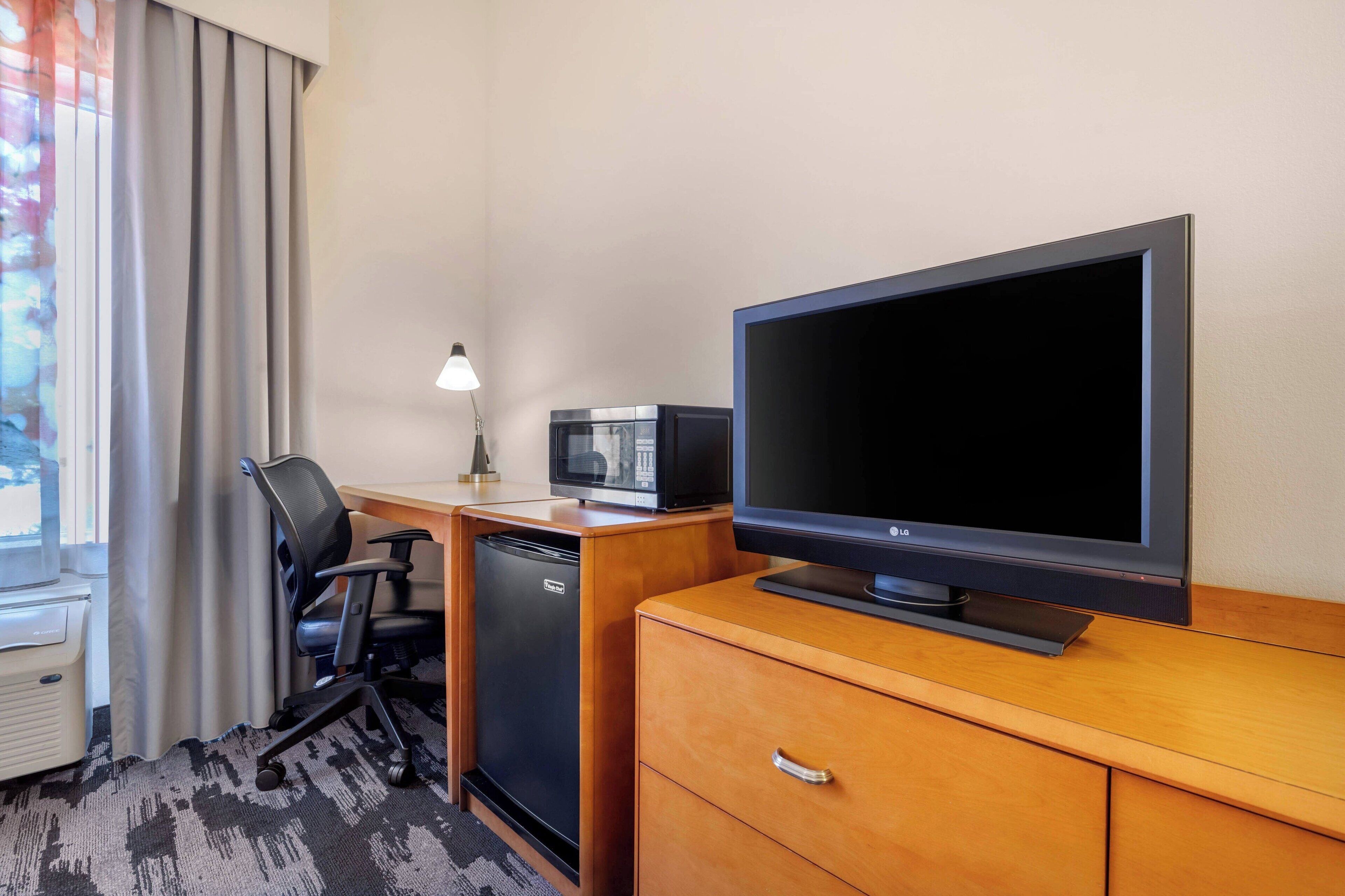 A hotel room with a wooden desk, a flat-screen TV, a microwave, a black office chair, a small refrigerator, and a lamp.