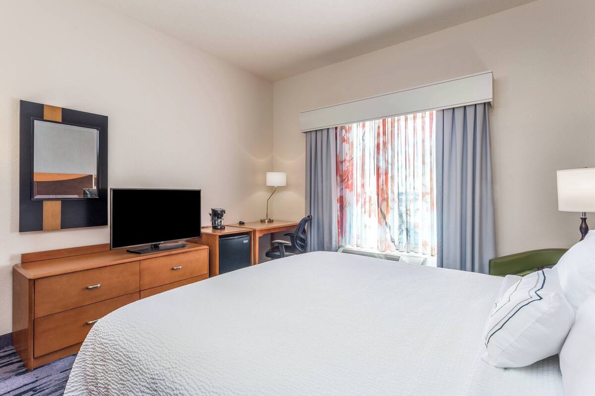 A hotel room with a large bed, a TV, a desk with a chair, a lamp, and a window with curtains.