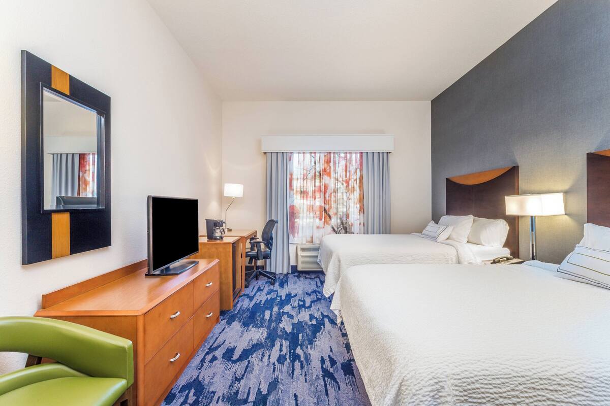 A hotel room with a large bed, a desk with a flat-screen TV, a chair, a desk lamp, and a window with curtains.
