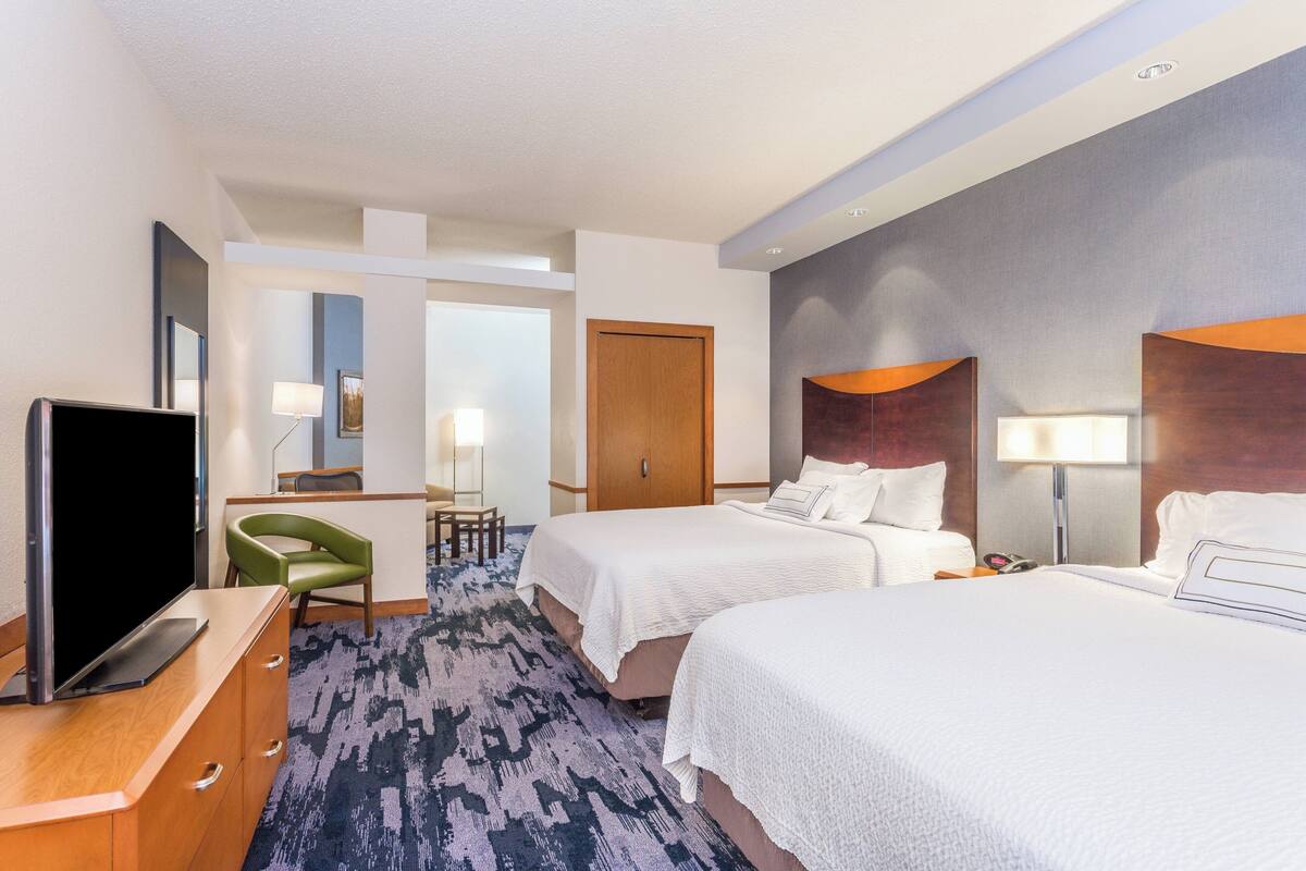 A hotel room with two beds, a desk with a flat-screen TV, a chair, and a carpeted floor.