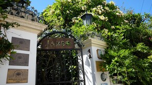 Ein schmiedeeisernes Tor mit dem Schriftzug „Jambo“ darüber, umgeben von üppigem Grün und blühenden Pflanzen.