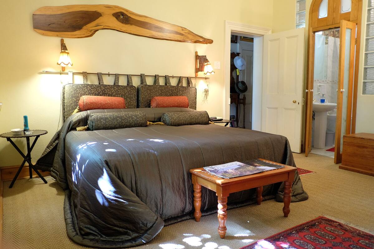 Une chambre à coucher avec un grand lit, deux lampes fixées au mur, une tête de lit en bois, une petite table avec un magazine et une porte donnant sur une salle de bain.