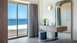 Une chambre d’hôtel moderne dotée d’une grande porte-fenêtre coulissante donnant sur la mer, d’une coiffeuse en bois avec un miroir et d’une petite plante en pot.