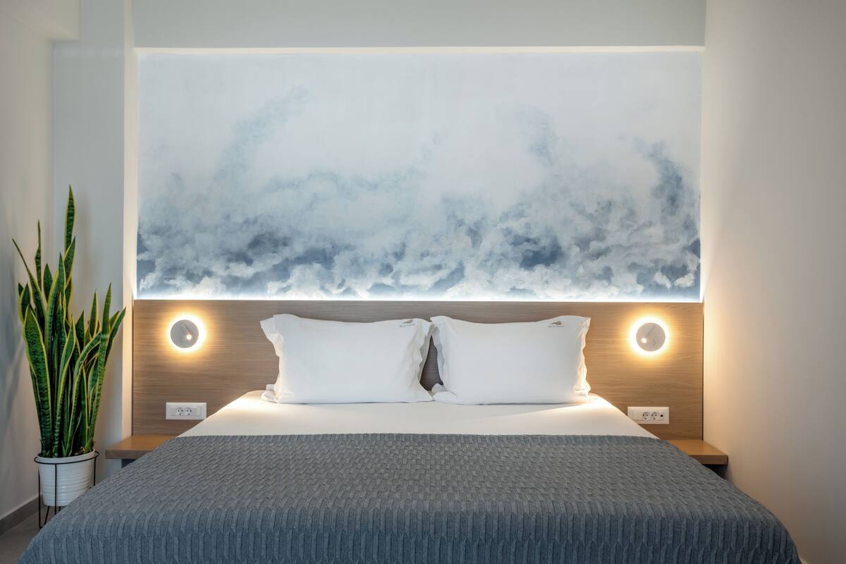 Habitación de hotel moderna con una cama grande, una lámpara de pared, una planta en maceta y una obra de arte con motivos de nubes sobre la cama.