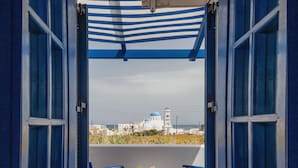 Un balcon avec deux chaises bleues et une table, donnant sur une ville côtière aux bâtiments blancs et à l’église à coupole bleue.