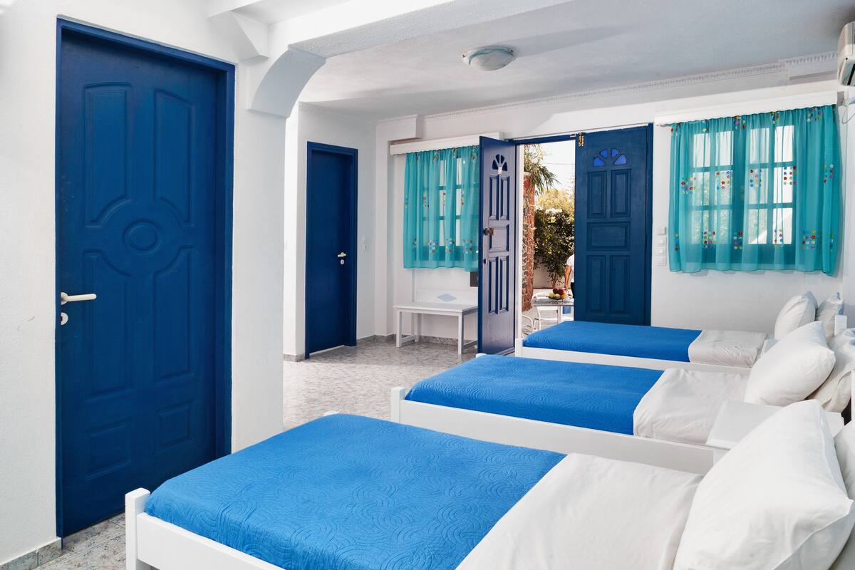 A room with two beds, a small table, and a chair. There are two doors, one blue and one white.