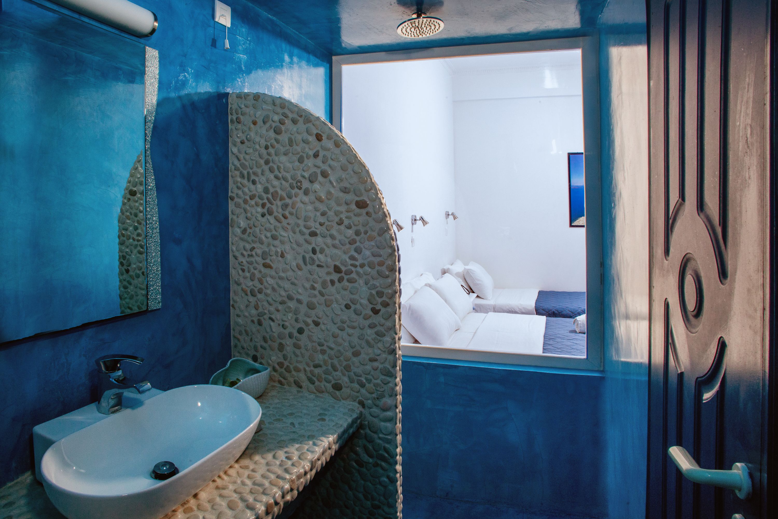Un baño con un lavamanos de textura de piedra, un espejo y vistas a un dormitorio con bañera y ventana.