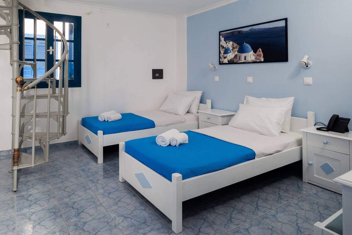 A hotel room with two beds, a small desk, a chair, a window with blue shutters, a painting of a coastal scene, and a security camera.