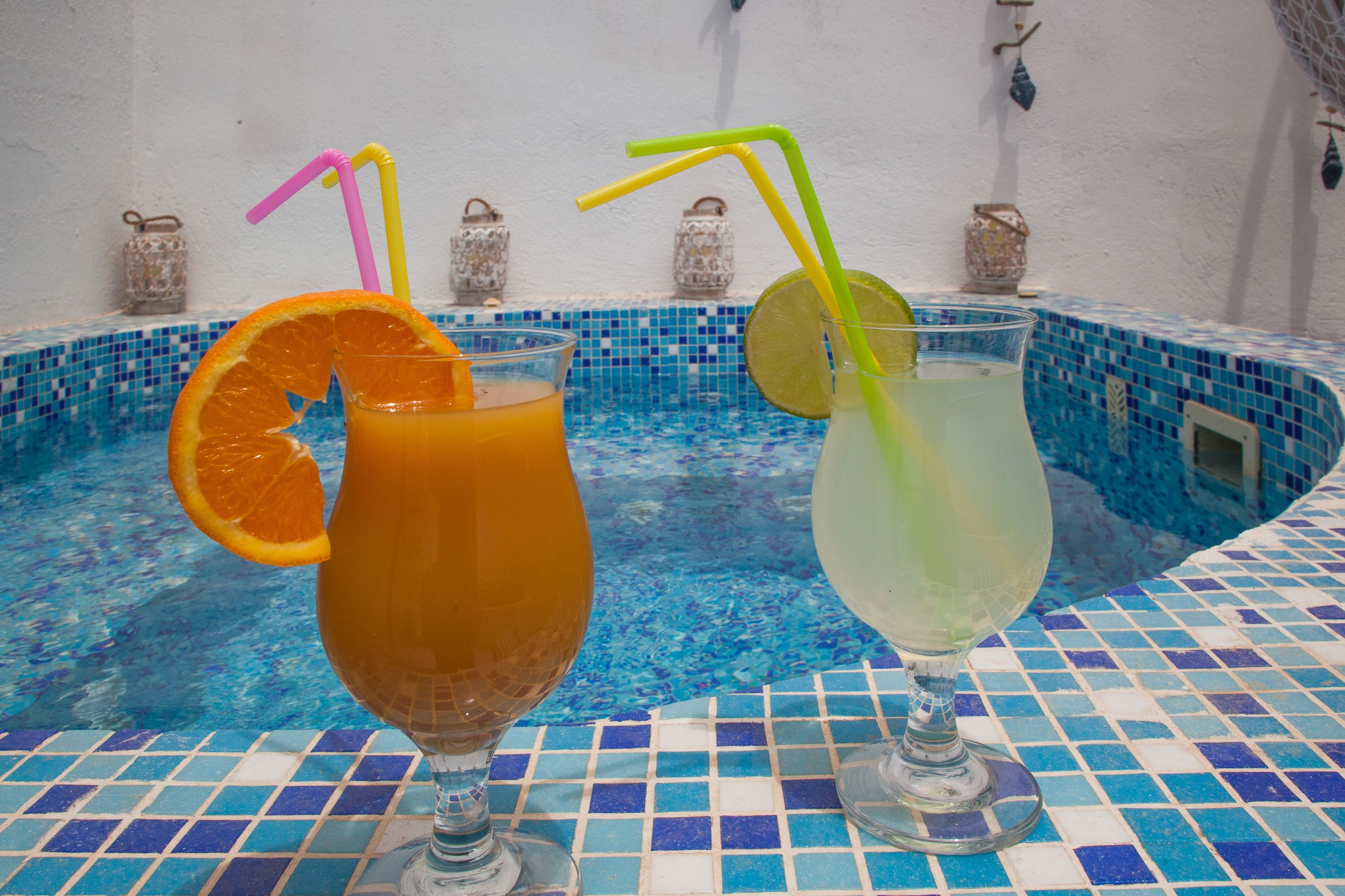 Two glasses of beverages with straws and slices of fruit on a tiled poolside table.