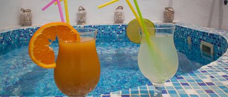 Two glasses of beverages with straws and slices of fruit on a tiled poolside table.