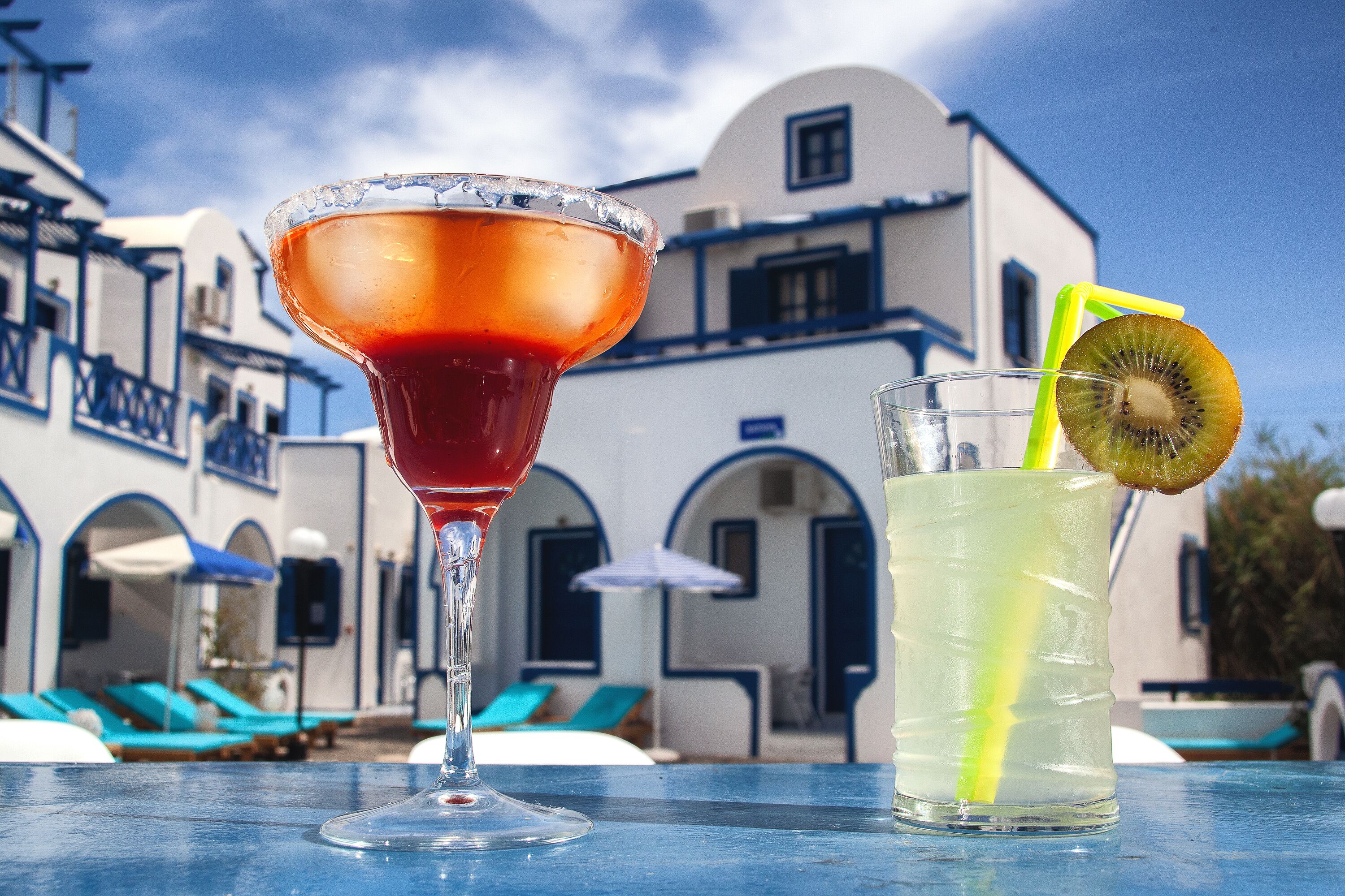 A table with two drinks, one in a martini glass and one in a highball glass, with a building in the background.
