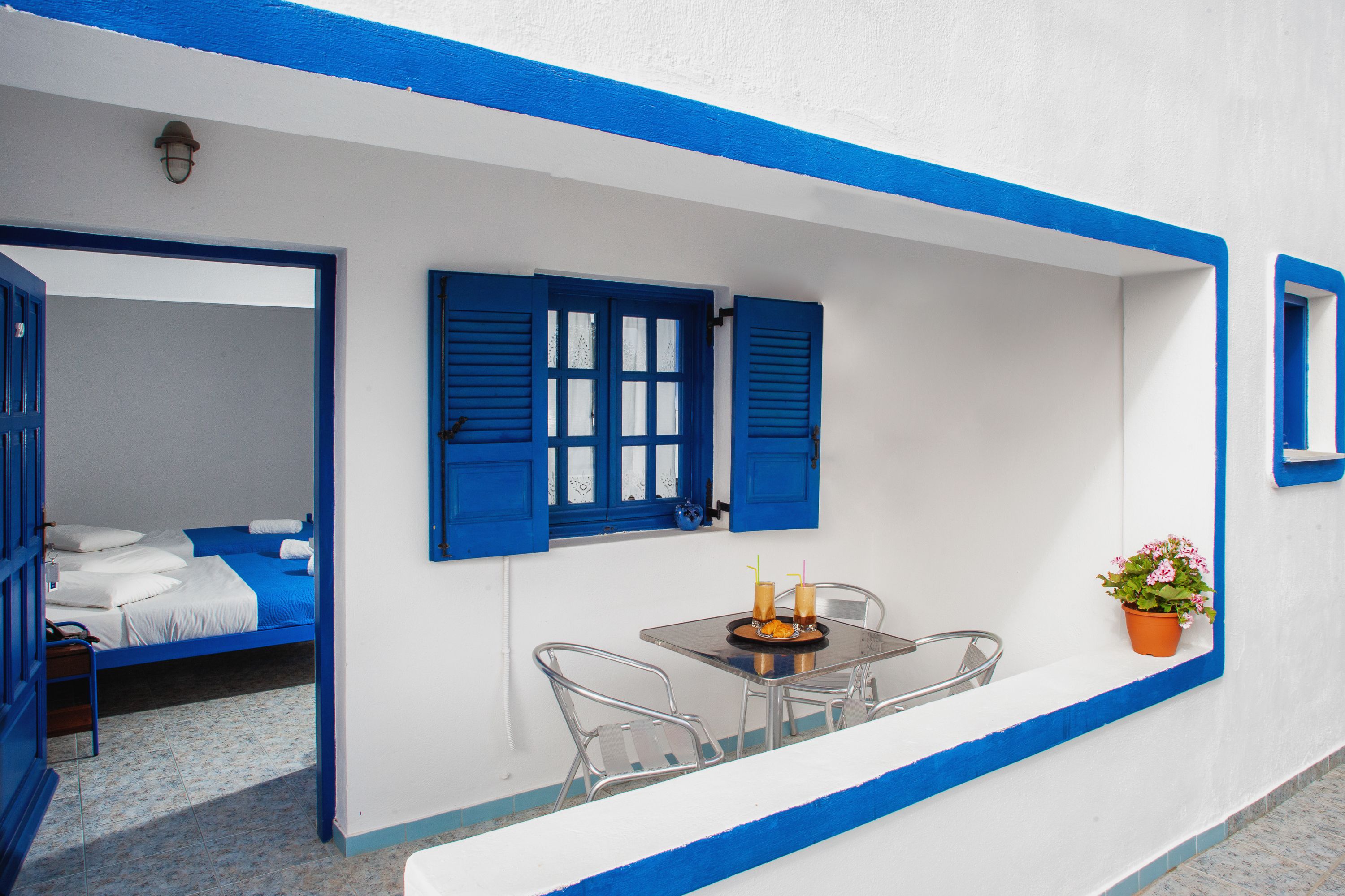 Een kamer met een bed, een tafel met stoelen, een raam met blauwe luiken en een kamerplant.