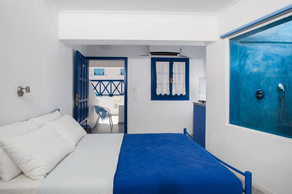 A bedroom with a bed, a window with curtains, a blue door, and a view of the outside.