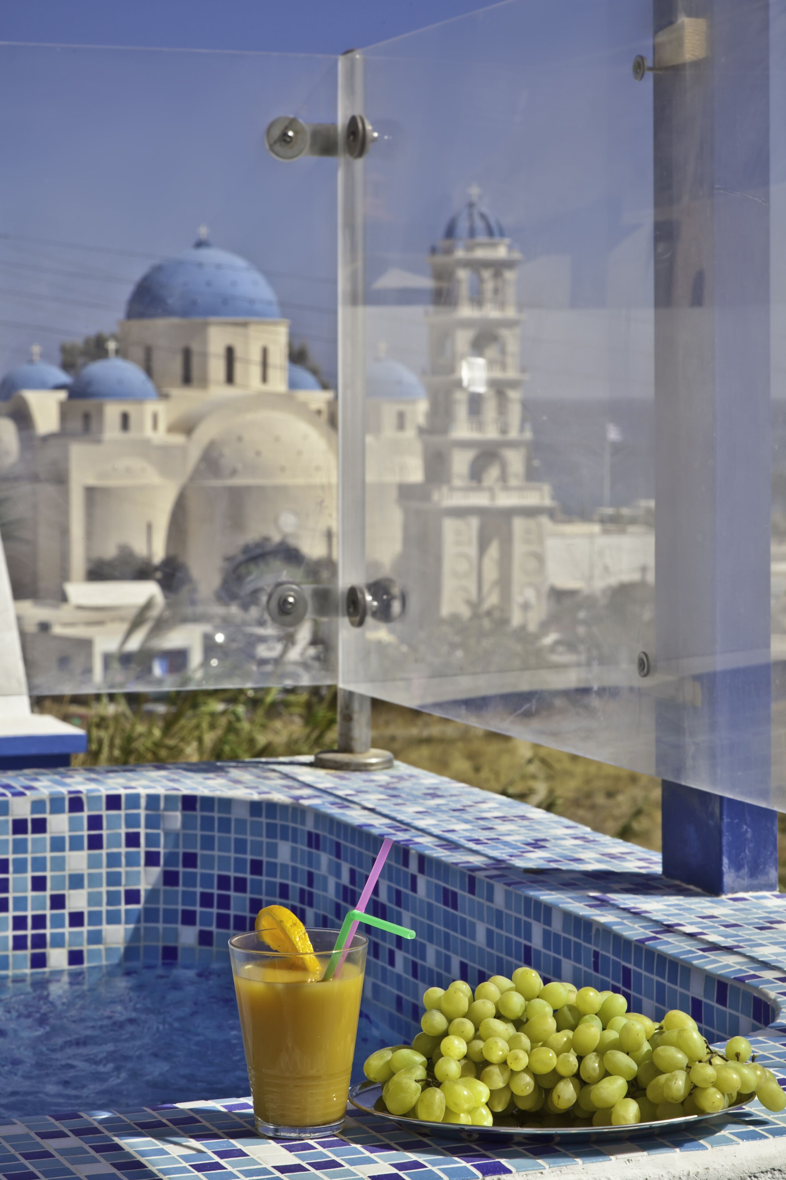 A table with a glass of orange juice and a plate of green grapes, a tiled blue and white tablecloth, and a view of a building with domed blue roofs.