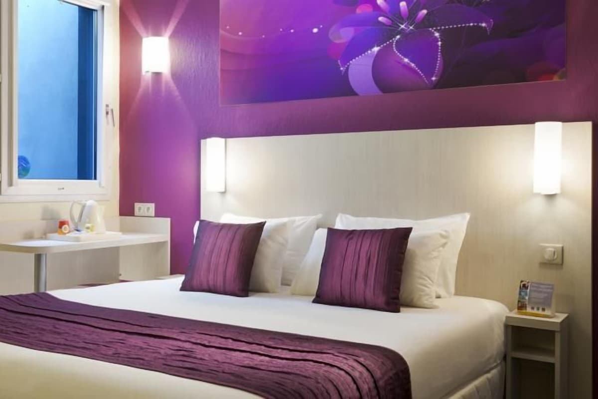A hotel room with a large bed, a bedside table, a nightstand, a window, and a wall-mounted light fixture.