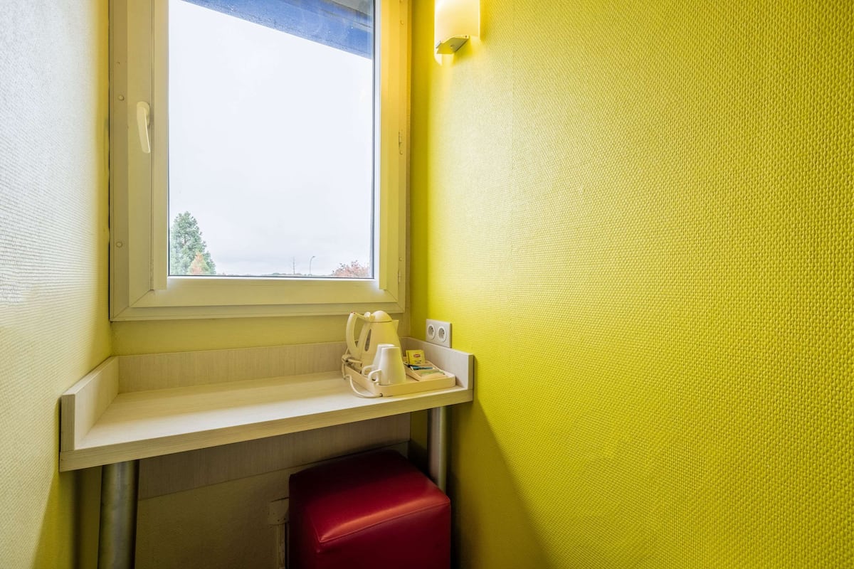 A small room with a window, a red stool, a shelf with a kettle, cups, and a tray, and a yellow textured wall.