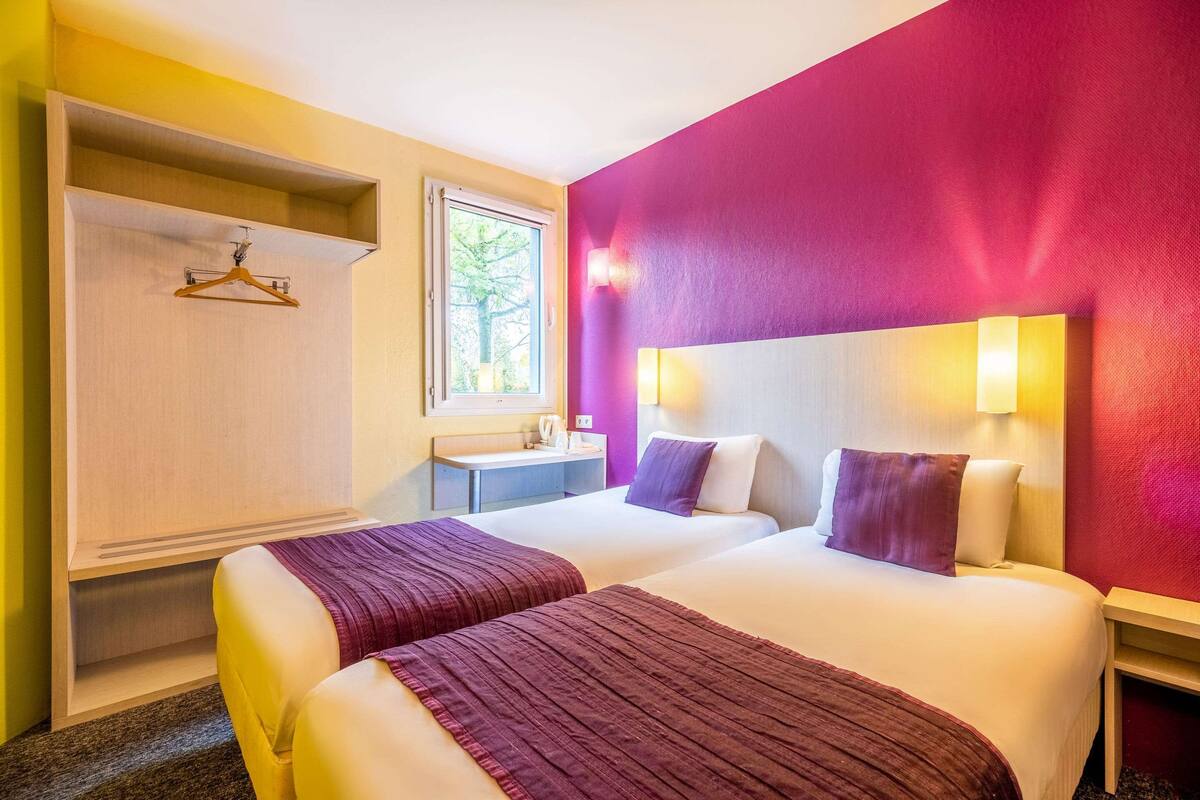 A hotel room with two beds, a wardrobe, a small desk, and a window.