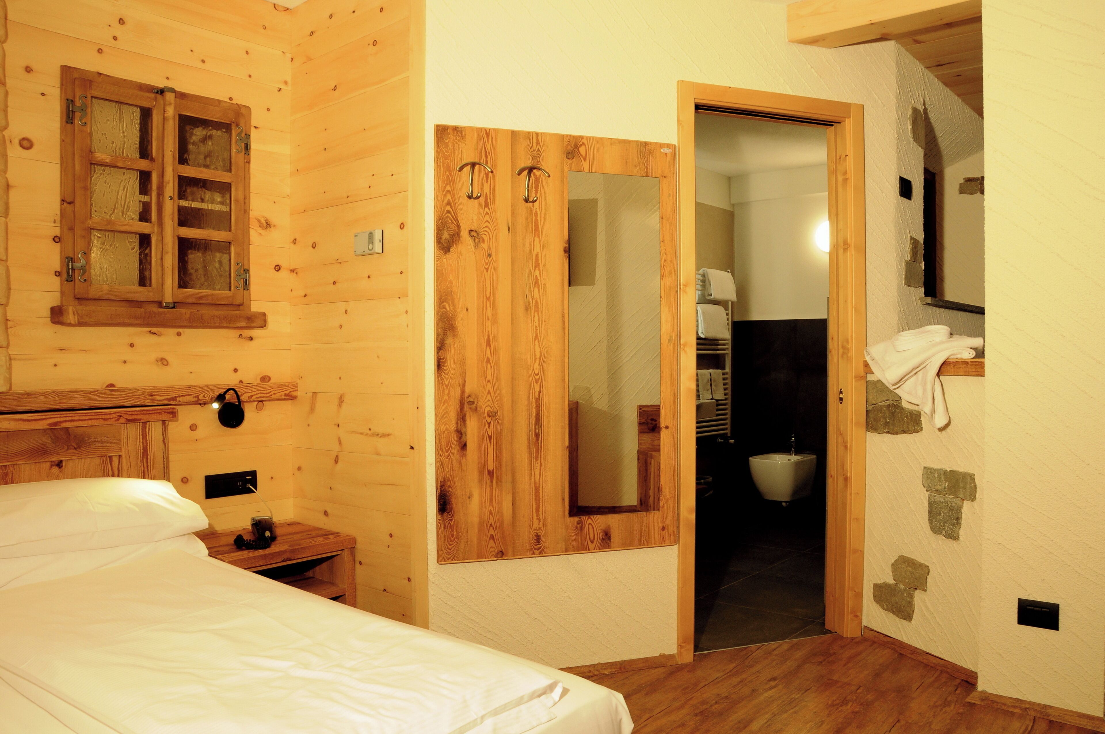 Habitació de fusta amb un llit, un armari de fusta, un mirall i un bany visible a través d'una porta oberta.