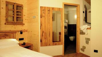 Una stanza in legno con un letto, un armadio in legno, uno specchio e un bagno parzialmente visibile da una porta aperta.