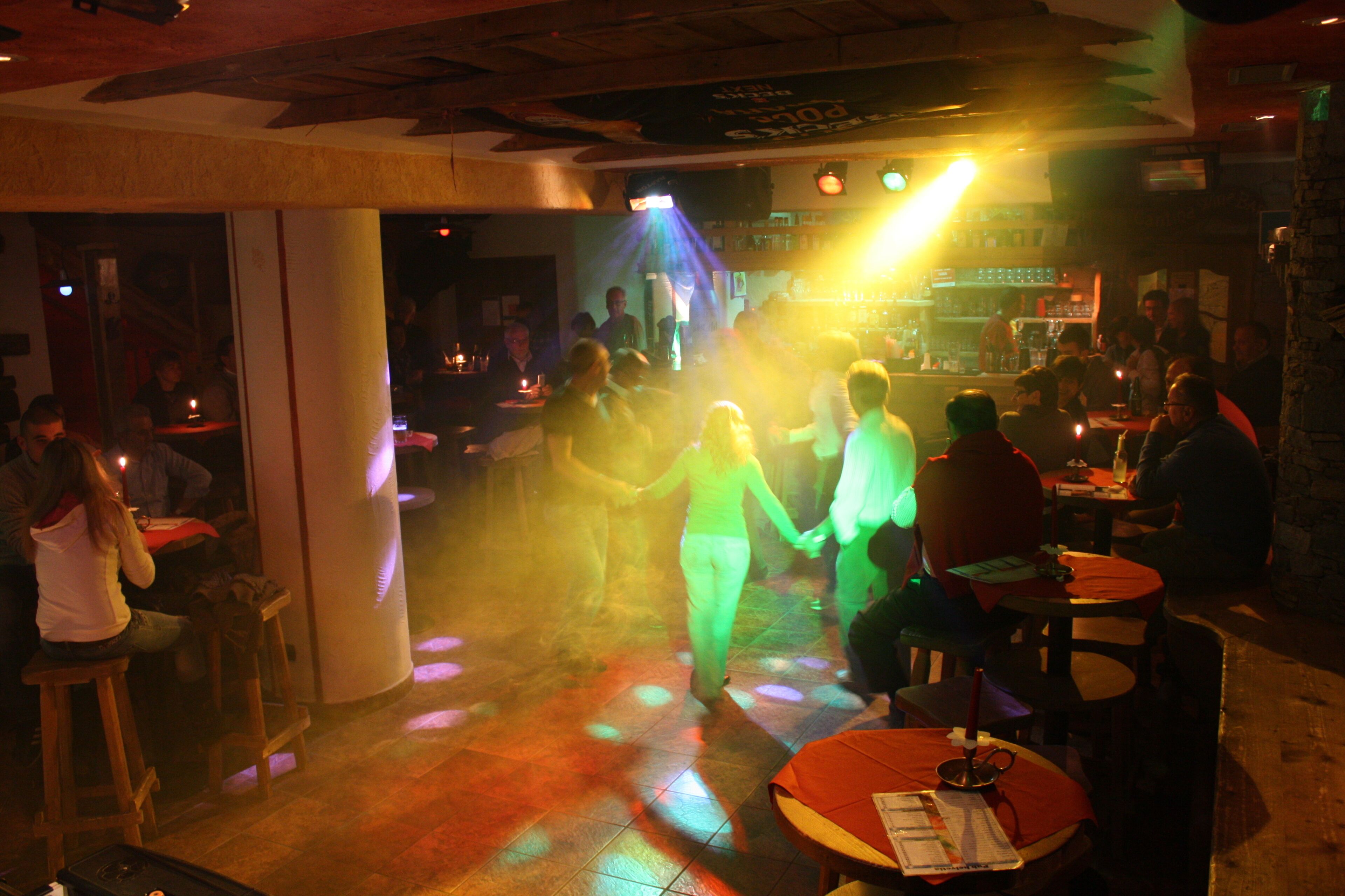 A bar with tables and stools, people dancing, and a stage with a DJ.