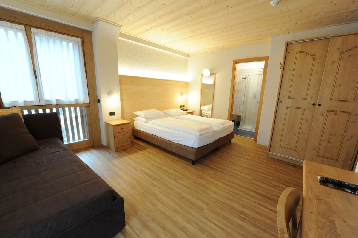 A room with a bed, a sofa, a wooden wardrobe, a bedside table, and a mirror.