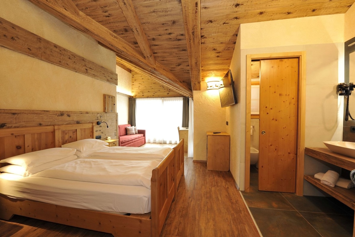 A bedroom with a wooden bed, a small bedside table, a red chair, a TV mounted on the wall, and a wooden door leading to a bathroom.