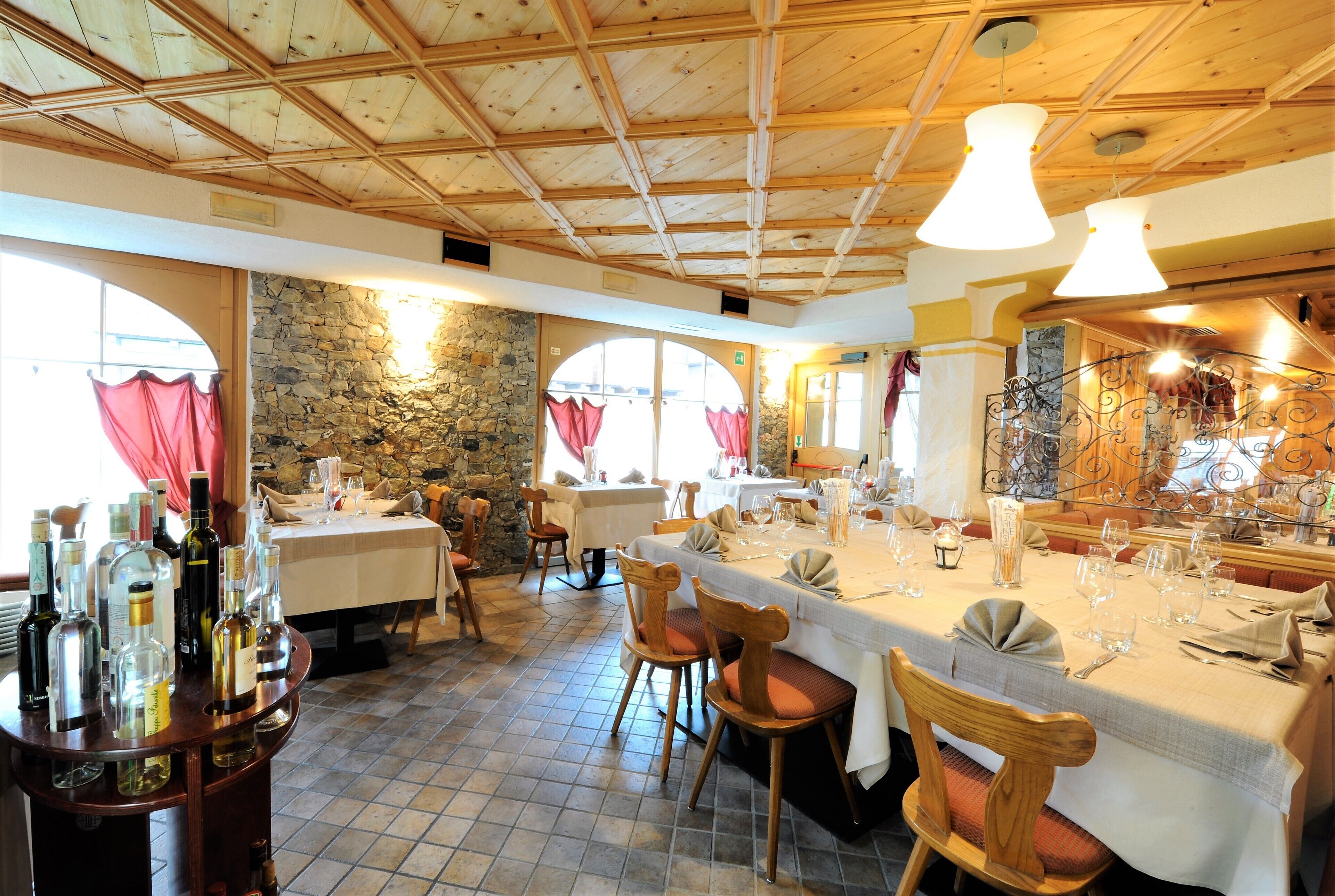 A restaurant with wooden ceiling beams, stone walls, and tiled flooring. Tables are set with wine glasses and bottles.
