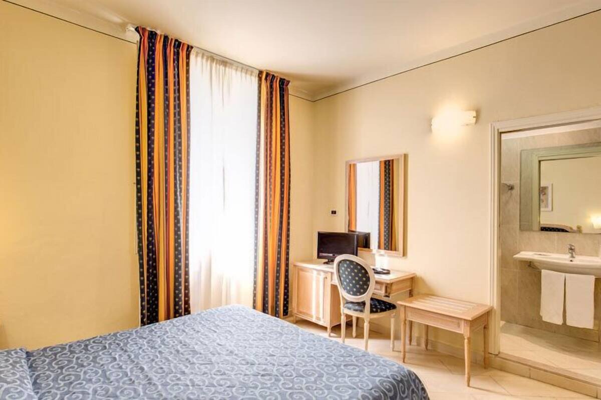 A hotel room with a bed, a desk with a computer, a chair, a mirror, and a bathroom with a sink and towel rack.