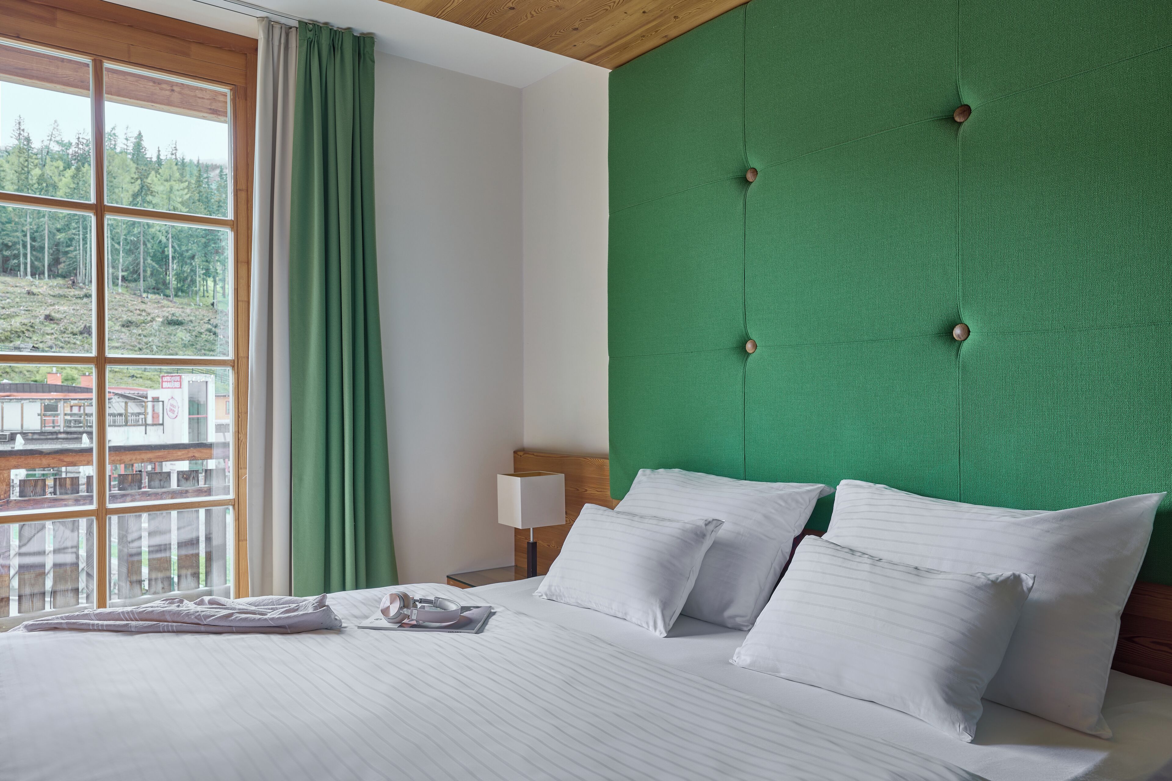 Ein ordentlich bezogenes Bett mit weißen Leinen und Kissen, ein mit Grün bezogener Kopfteil, eine Nachttischlampe und ein Fenster mit Blick auf eine bewaldete Landschaft.