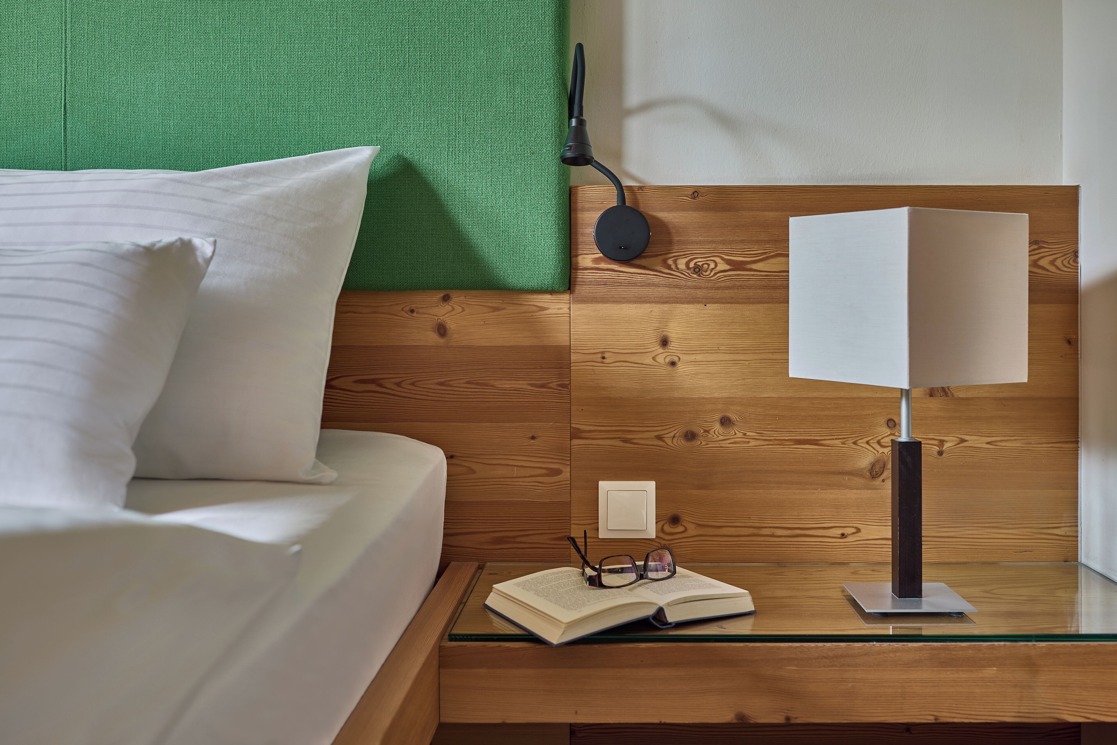 Una cama bien hecha con ropa de cama blanca, una mesita de noche con lámpara y una pared verde con textura.