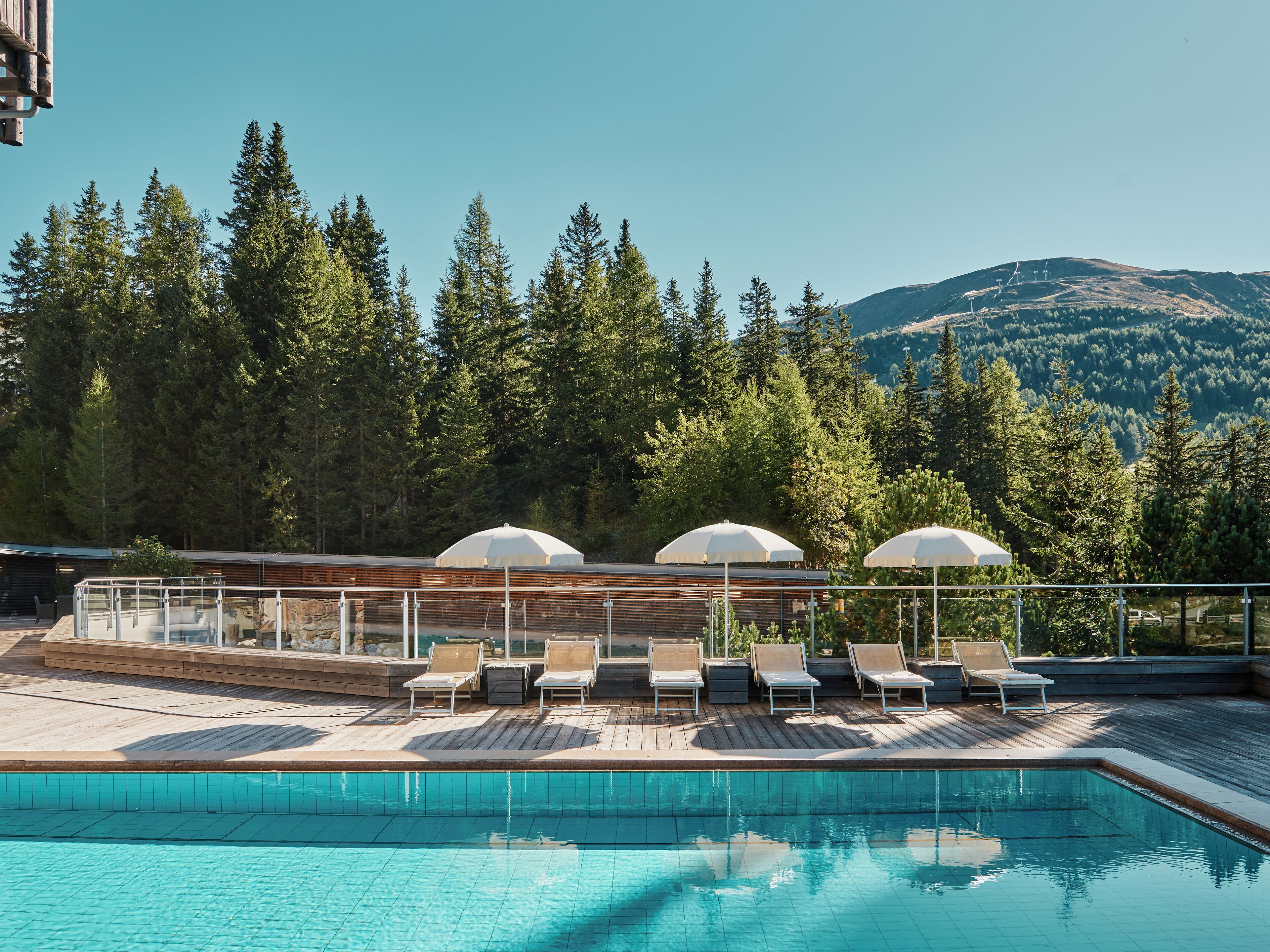 Un área de piscina con sillones de descanso y sombrillas, rodeada por una terraza de madera y con vistas a una cadena montañosa boscosa.