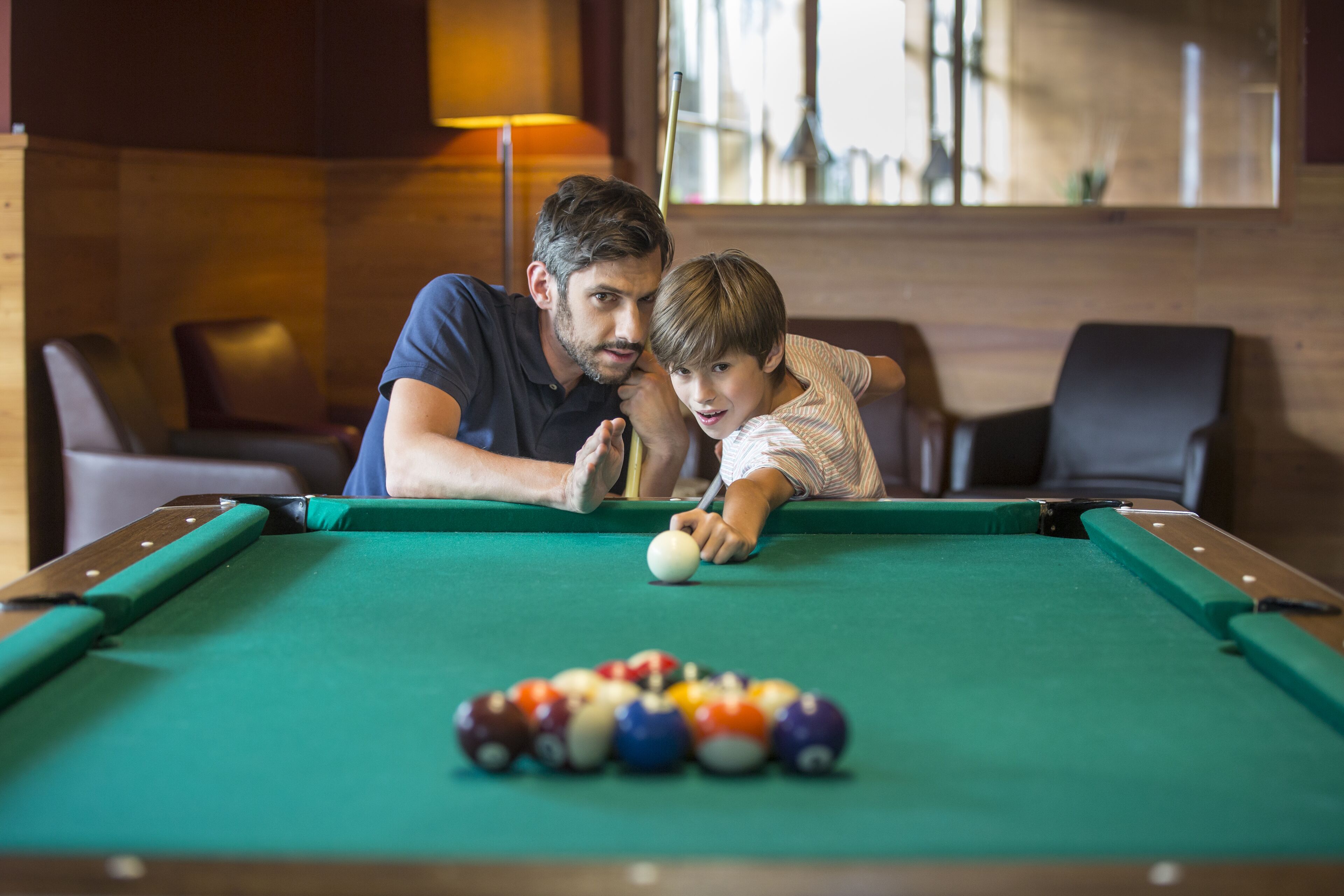A pool table with a cue ball and a rack of billiard balls, a wooden wall, and a lamp in the background.