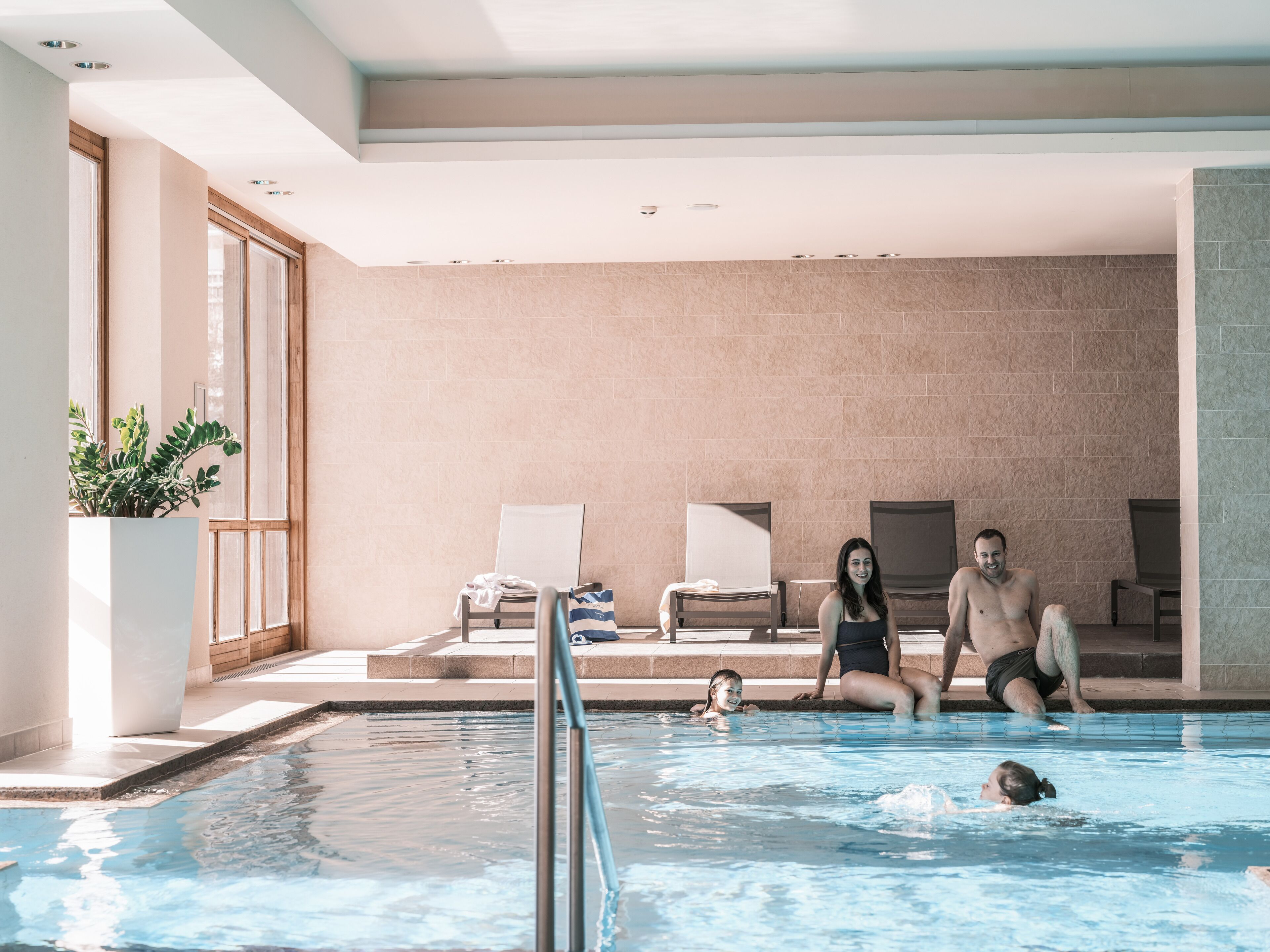 An indoor swimming pool with a tiled wall, a potted plant, and lounge chairs.