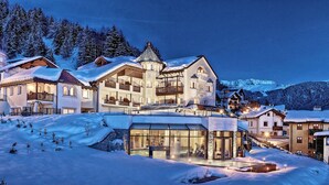 Un paese montano imbiancato dalla neve, con numerosi edifici, tra cui un albergo con balconi e ampie finestre, sullo sfondo di montagne innevate.
