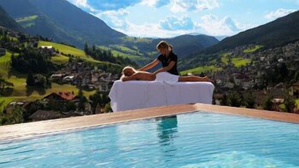 Una persona si rilassa su una poltrona bianca a bordo piscina, con un paesaggio montuoso sullo sfondo.