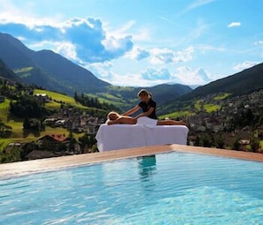 A person relaxing on a white lounge chair by a pool with a mountainous landscape in the background. - Alpenheim Charming & Spa Hotel (Ortisei)