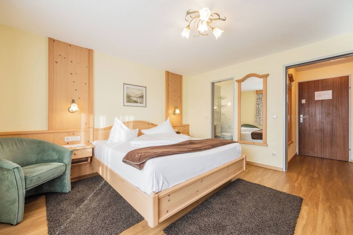 A bedroom with a wooden bed, a green armchair, a bedside table, a mirror, and a wooden wardrobe.