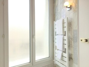 A bathroom with a frosted glass door, a towel rack with multiple towels, a wall-mounted light fixture, and a marble-like backsplash.