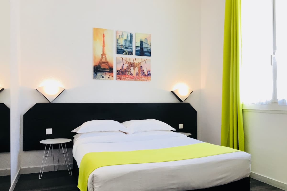A hotel room with a bed, a small table, two wall-mounted lamps, and a window with curtains.