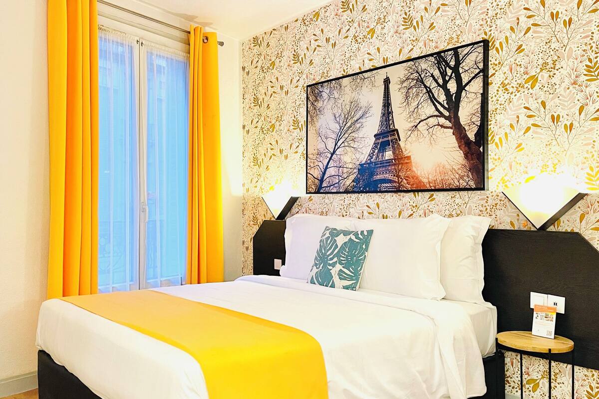 A hotel room with a bed, a window with yellow curtains, a wall-mounted TV, and a painting of the Eiffel Tower.
