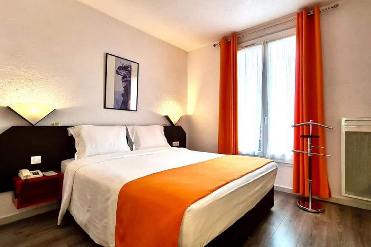 A hotel room with a bed, bedside tables, a desk, a chair, a window with curtains, and a wall-mounted air conditioning unit.