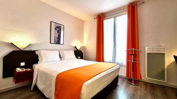 A hotel room with a bed, bedside tables, a desk, a chair, a window with curtains, and a wall-mounted air conditioning unit.