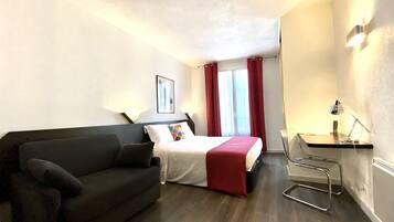 A compact hotel room with a bed, a desk with a chair, a sofa, and a window with curtains.
