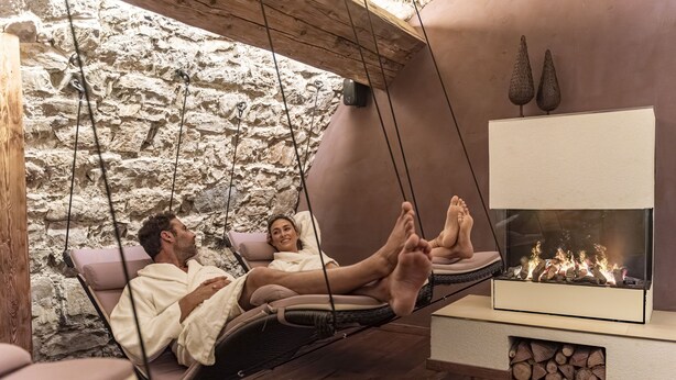 A couple relaxing in a hammock by a fireplace in a rustic stone-walled room.