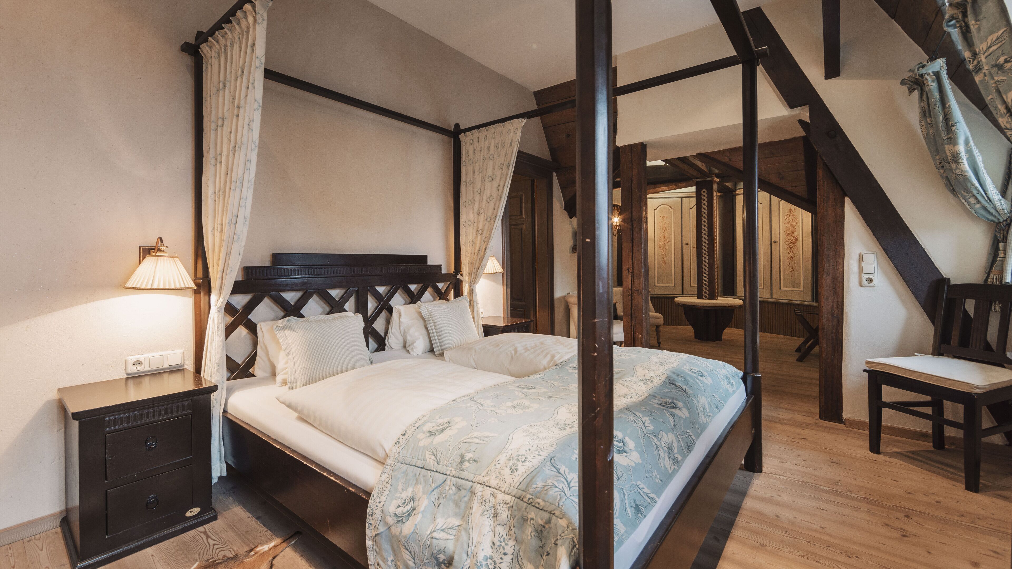 Ein Bett mit Baldachin, eine Blumendecke, ein Nachttisch mit Lampe und ein Holzstuhl in einem Zimmer mit Holzboden und schrägen Wänden.