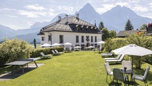 Un hotel con un césped bien cuidado, mobiliario exterior con sombrillas y montañas al fondo.